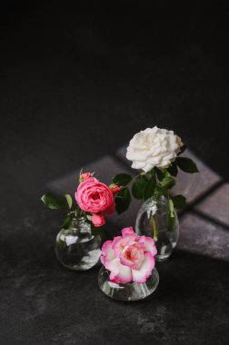 Koyu dokulu yüzeyde küçük cam vazolarda üç narin gül, yumuşak ışık.