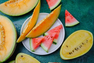 Taze kavun dilimleri ve karpuz dilimleri desenli yeşil bir zemin üzerine dizilmiş renkli meyve çeşitleri sunar..