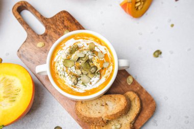 Kremalı balkabağı çorbası ve tahtanın üzerinde ekmek dilimleri olan beyaz kasede tohumlar.