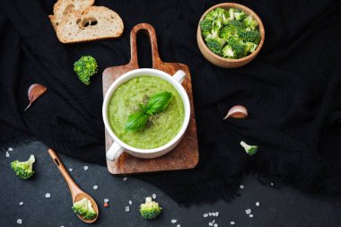 Brokoli çiçekli kremalı yeşil çorba ve ahşap bir tahtada taze fesleğen garnitürü, ekmek ve sarımsak ile servis edilir.