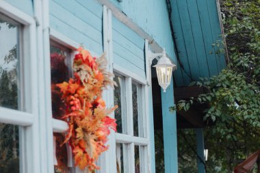 Soluk mavi ev penceresinde sonbahar yaprağı çelengi ve sıcak açık hava aydınlatma armatürü.