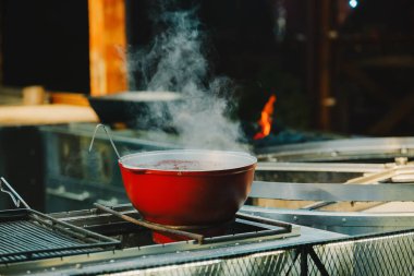 Metal tezgahın üzerinde sıcak sıvı ile buharlaşan büyük kırmızı tencere. Parlak alevlerle dolu açık hava sahnesi.