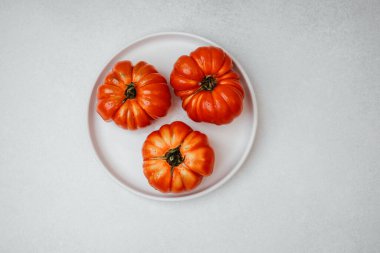 Açık gri bir yüzey üzerinde beyaz bir tabağa su damlatan üç olgun kırmızı aile yadigarı domates..