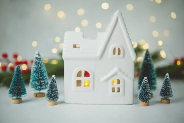 Beyaz seramik ev minyatür çam ağaçları ve yumuşak bokeh ışıklarıyla çevrili.