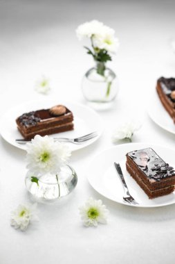 Beyaz tabakta çikolatalı pasta dilimi ve beyaz çiçekler ve masada çatallar.