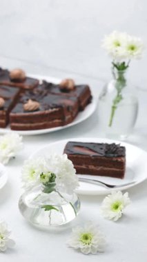 Temiz bir masadaki cam vazolarda beyaz çiçekli zengin çikolatalı pasta dilimleri.