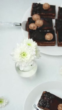 Üzerinde krema olan çikolatalı kekler beyaz tabaklarda servis ediliyor ve yanında beyaz çiçekler var.