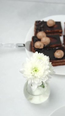 Beyaz tabakta kremalı çikolatalı kekler, masada vazoda beyaz çiçeklerle süslenmiş.