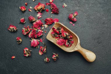 Koyu bir yüzeyde ahşap bir kepçe içinde kurumuş gül tomurcukları, dağınık yapraklar.