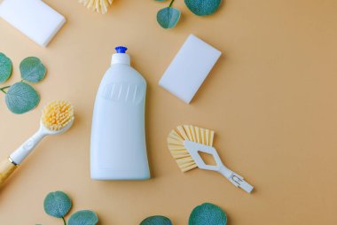 Temizlik fırçası ve süngerli beyaz şişe, doğal yeşil yapraklar, açık kahverengi arka planda basit temizlik malzemeleri..