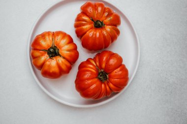 Açık gri bir yüzey üzerinde beyaz bir tabağa su damlatan üç olgun kırmızı aile yadigarı domates.