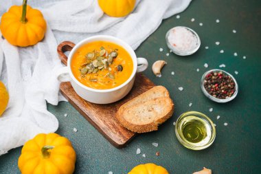 Bir kase kremalı balkabağı çorbası, üzerinde tohumlar, ekmek dilimleri ve kırsal bir panoda baharatlar..