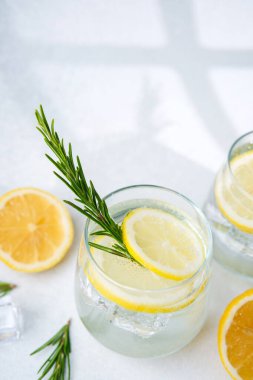 Bir bardak köpüklü limon suyu, biberiye filizi, buz küpü ve taze limon. Yarısı beyaz yüzeyde parlak güneş ışığı altında.