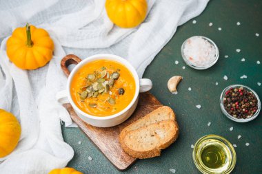 Beyaz kasede sağlıklı balkabağı çorbası. Arkaplanda tohum, ekmek, baharat ve yağ var.