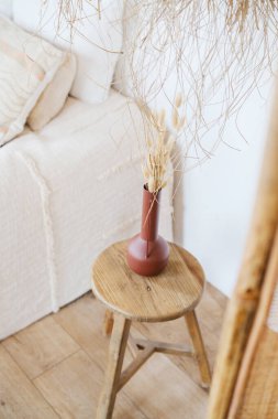 Yumuşak yatak odasının yanındaki ahşap taburede kuru çimen bulunan seramik vazo.
