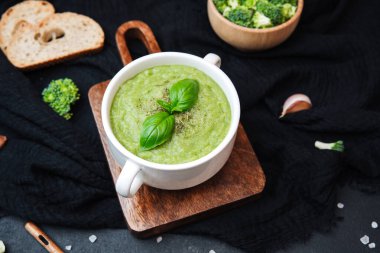 Fesleğen yaprağı ve kurutulmuş otlarla süslenmiş beyaz kâse içinde yeşil brokoli çorbası, ekmekle servis edilir.