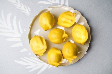 Taze sarı limonlar bej plaka üzerinde sergileniyor. Yaprak şeklinde gölgeler, gri arka planda güneş ışığıyla dökülüyor.