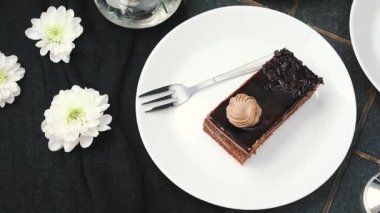 Beyaz tabakta çikolatalı pasta dilimi, çatallı ve siyah kumaşlı beyaz çiçekler.