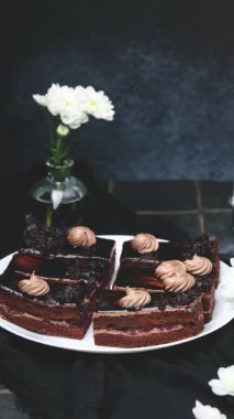 Beyaz çiçeklerin yanındaki beyaz tabakta kremalı çikolata katmanlı kekler.