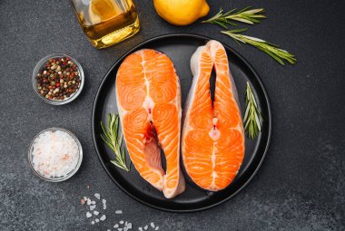 Salmon fillets laid on a black plate with rosemary, olive oil, lemon, and spices nearby.