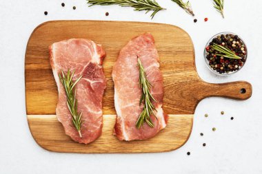 İki çiğ domuz pirzolası, etrafına biberiye ve baharat konmuş ahşap bir tahtaya..