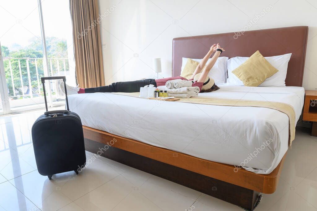 Portrait of tourist woman lying on the bed, stretching her hands with her luggage in hotel bedroom after check-in. Conceptual of travel and vacations.