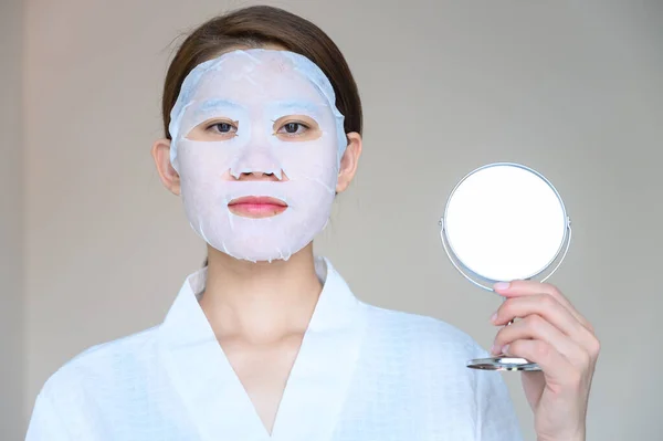 Portrait of young woman holding a mirror while applying facial mask for enhance her skin. Facial mask is a creamy or thick pasted mask applied to clean or smoothen the face.
