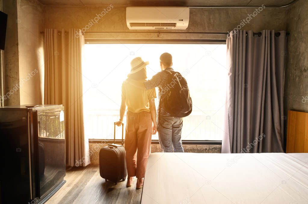 Portrait of young couple tourist standing nearly window, looking to beautiful view outside in hotel/resort bedroom after check-in. Conceptual of couple travel and vacation in their honeymoon period.
