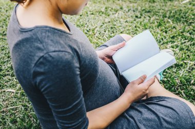 Genç hamile bir kadının bahçede oturup kitap okurken çekilmiş görüntüsü (boş alanı olan bir kitap). Hamile kadın yaşam tarzı kavramı.