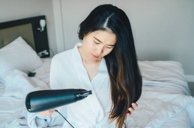 Saç kurutma makinesi kullanan Asyalı genç bir kadının portresi sıcak havayı üfleyerek yatak odasındaki saçı kurutmak için suyun buharlaşmasını hızlandırıyor. Kadın yaşam tarzı kavramı.
