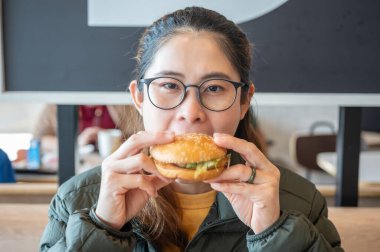 Hamburger yerken genç Asyalı bir kadının görüntüsü. Hamburger, ekmeğin üzerinde pişmiş köfte ya da rulo köfteden oluşan bir sandviçtir..