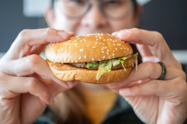 Yemeden önce elinde hamburgerle gezen Asyalı genç bir kadın görüntüsü. Hamburger, ekmeğin üzerinde pişmiş köfte ya da rulo köfteden oluşan bir sandviçtir..