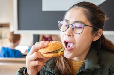 Genç Asyalı bir kadının hamburgeri yemek için ağzını açtığı görüntüler. Hamburger, ekmeğin üzerinde pişmiş köfte ya da rulo köfteden oluşan bir sandviçtir..