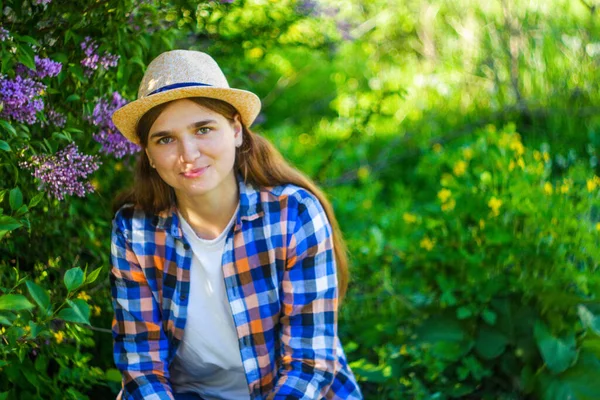 Rozostřit mladou letní ženu. Krásná žena v kostkované košili a klobouku s šeříkovou partou a loukou květin na jarní zahradě, portrét mladých žen. Rozostřeno — Stock fotografie