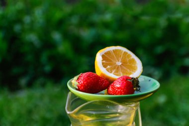 Limonlu Defocus çay tabağı ve doğanın yeşil arka planında cam bir sürahi limonata üzerinde duran iki çilek. Yaz meyveleri. Odaklanamıyorum.