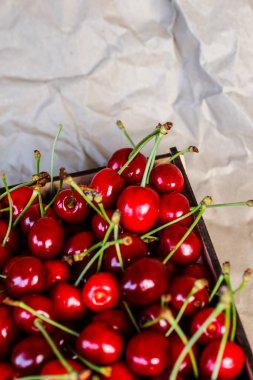 Cropped wooden box, crape of sweet red cherries with tail and leaves on craft yellow wrinkled paper background. Summer fruits and berries. Harvest and crop concept. Organic food. Top view. Diagonal.
