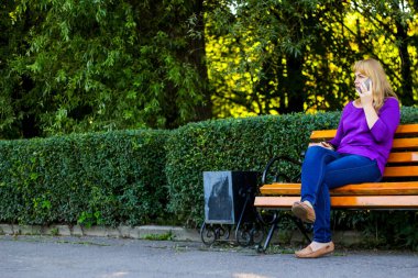 Defocus caucasian blond woman talking, speaking on the phone outside, outdoor. 40s years old woman in purple blouse in park on bench. Adult female using cellphone. Copy space. Out of focus.