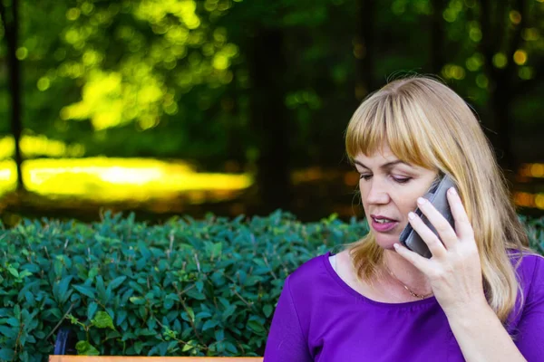 Defocus elegance caucasian blond woman talking, speaking on the phone outside, outdoor. 40s years old woman in purple blouse in park on bench. Adult women using telephone. Copy space. Out of focus.