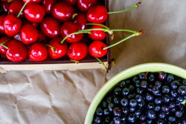 Cropped bowl of blueberries and box, crape of red sweet cherries with tail on craft wrinkled old paper background. Harvest and crop concept. Organic food. Top view. Summer fruits. Flatly. Still life.