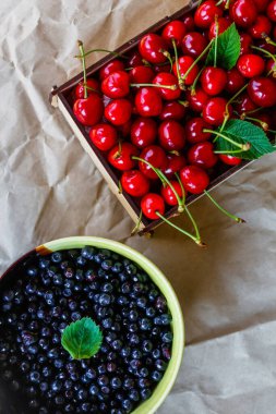 Kırpılmış yaban mersini kasesi ve kutu, buruşuk kağıt arka planda kuyruğu olan kırmızı tatlı kirazlar. Dikey. Organik yiyecekler. Üst Manzara. Yaz meyveleri ve meyveler. Dümdüz. Durgun hayat