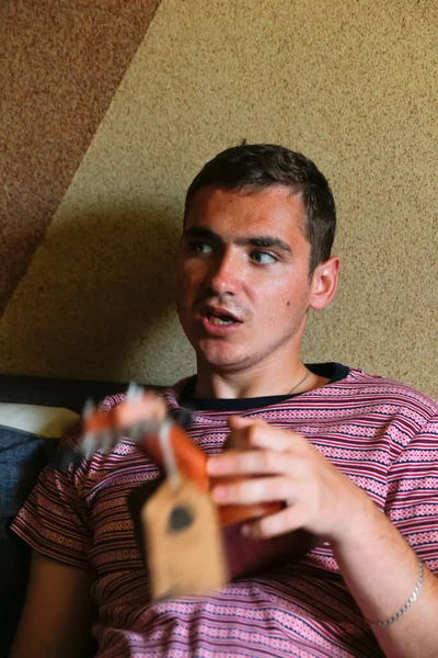 Hombre joven de 20 años confundido con grandes ojos sosteniendo una pequeña guitarra ukelele y hablando. Sorprendido tipo de pelo castaño caucásico en el interior. Hombres desconcertados. Vertical — Foto de Stock