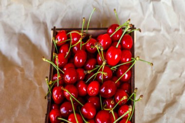 Kırpılmış kutu, buruşuk kağıt arka planda kuyruğu olan koyu kırmızı kirazlar. Yaz meyveleri ve meyveler. Hasat ve hasat konsepti. Organik yiyecekler. Üst Manzara. Yakın plan..