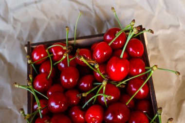 Kırpılmış kutu, kırmızı kiraz çatırtısı kuyruklu ve yaprakları buruşuk kağıt arka planda. Yaz meyveleri ve meyveler. Hasat ve hasat konsepti. Organik yiyecekler. Üst Manzara. Çapraz. Yakın plan..