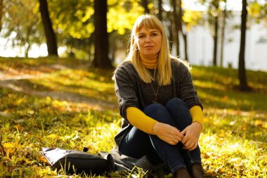 Sarı sonbahar parkında oturan 40 'lı yaşlardaki güzel sarışın kadını defocus. Mutlu güzel bayan. Gri kazak giyen kadınlar, sarı balıkçı yaka, kolye, kot pantolon. Sonbahar parkı, yapraklar, gün batımı. Odaklanamıyorum.