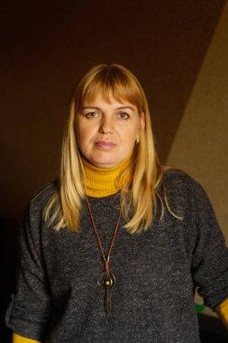 Evde dikilen 40 'lı yaşlardaki olgun sarışın kadının defocus' u. Mutlu güzel bayan. Gri kazak giyen kadınlar, sarı balıkçı yaka, kolye. Dikey portre. Odaklanamıyorum.