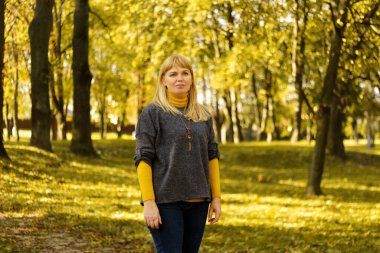 Sarı sonbahar parkında duran 40 'lı yaşlarda sarışın bir kadın. Mutlu güzel bayan. Gri kazak giyen kadınlar, sarı balıkçı yaka, kolye, kot pantolon. Yapraklarla dolu sonbahar parkı. Odaklanamıyorum.