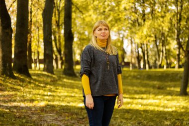 Sarı sonbahar parkında duran ciddi sarışın 40 'lı yaşlardaki Defocus. Mutlu güzel bayan. Gri kazak giyen kadınlar, sarı balıkçı yaka, kolye, kot pantolon. Yapraklarla dolu sonbahar parkı. Odaklanamıyorum.