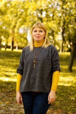 Sarı sonbahar parkında gülümseyen 40 'lı yaşlardaki sarışın kadın. Mutlu güzel bayan. Gri kazak giyen kadınlar, sarı balıkçı yaka, kolye, kot pantolon. Sonbahar parkı, yapraklar. Dikey. Odaklanamıyorum.