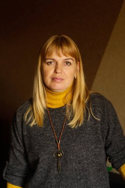 Evde dikilen 40 'lı yaşlardaki olgun sarışın kadının defocus' u. Mutlu güzel bayan. Gri kazak giyen kadınlar, sarı balıkçı yaka, kolye. Dikey portre. Odaklanamıyorum.