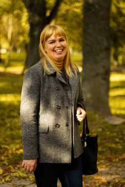 Sarı sonbahar parkında gülümseyen 40 'lı yaşlardaki sarışın kadın. Mutlu güzel bayan. Gri ceketli, kazaklı, sarı balıkçı yakalı kadınlar. Sonbahar parkı, yapraklar. Dikey portre. Odaklanamıyorum.